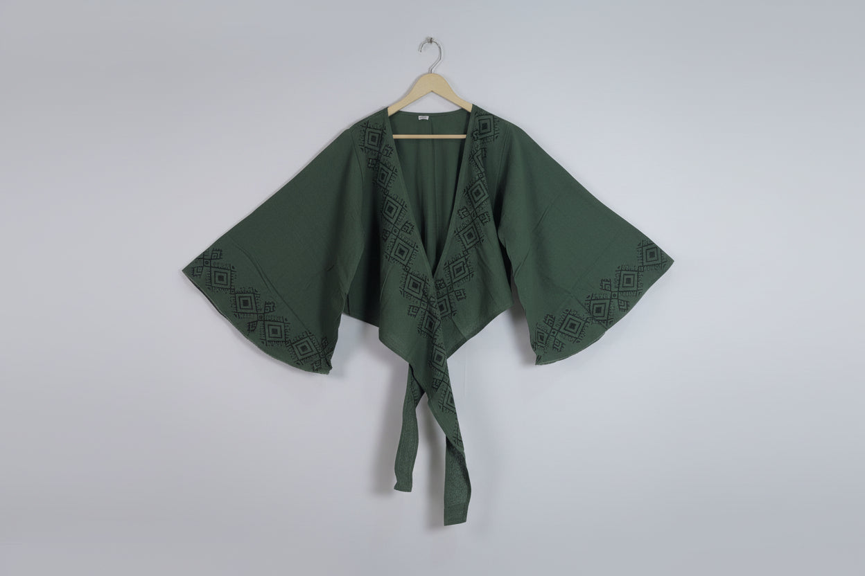 Green embroidered kimono-style top on a hanger against a white wall.